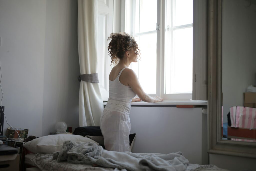 A woman in a bedroom gazes out the window, creating a calm and serene atmosphere.