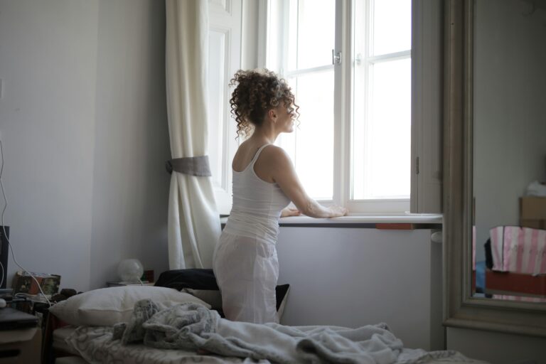 A woman in a bedroom gazes out the window, creating a calm and serene atmosphere.